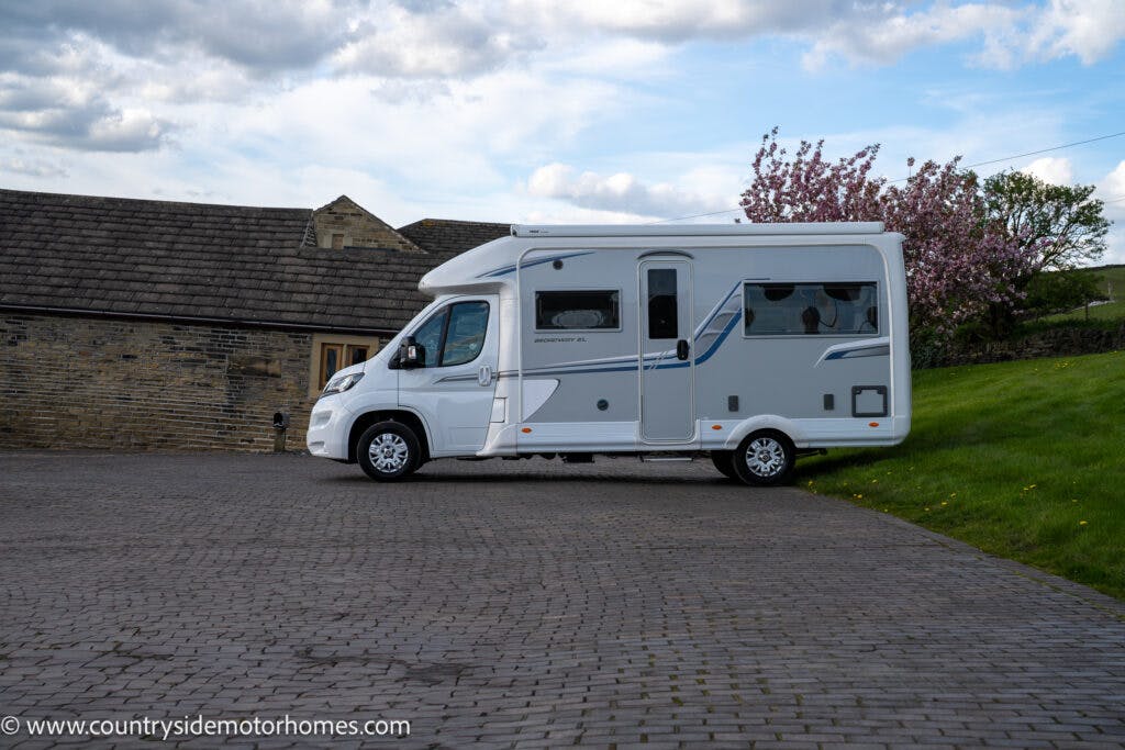 A white 2021 Auto-Sleepers Broadway EL motorhome is parked on a cobblestone driveway next to a stone building. A green grassy area with a blooming tree is visible in the background. The sky is partly cloudy. A website URL is printed at the bottom corner of the image.