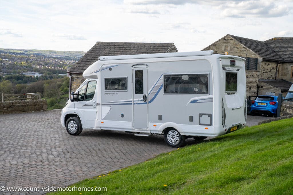 A 2021 Auto-Sleepers Broadway EL motorhome is parked on a paved driveway with a stone house and a blue car in the background. The landscape features rolling hills and scattered trees under a partly cloudy sky. The motorhome has blue and gray accents.