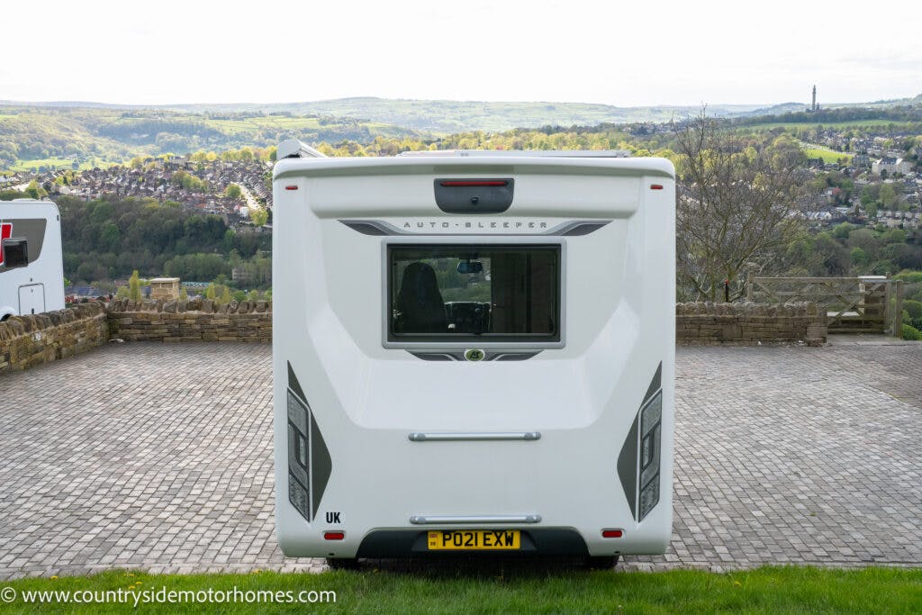A white 2021 Auto-Sleepers Broadway EL motorhome with the license plate "PO21 EXW" is parked on a brick driveway. The picturesque countryside with rolling hills and houses stretches out in the background. The website "www.countrysidemotorhomes.com" is printed at the bottom left of the image.