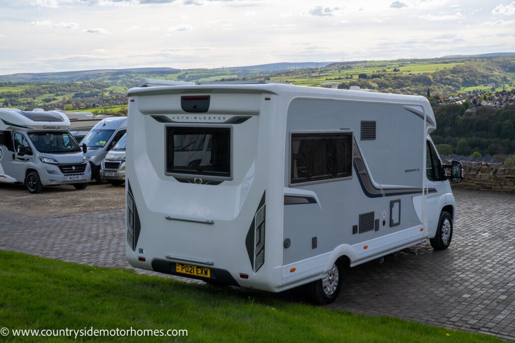 A white 2021 Auto-Sleepers Broadway EL motorhome is parked on a paved area with several other motorhomes in the background. The vehicle has a visible registration plate "PO21 EXN." The scene includes a lush landscape with green fields and hills under a partly cloudy sky.