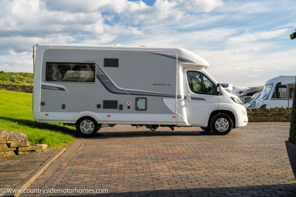 A 2021 Auto-Sleepers Broadway EL motorhome parked on a paved driveway with a grassy hill and a stone wall in the background. The motorhome features gray and blue detailing along the side. The sky is partly cloudy, and another motorhome is visible in the distance.