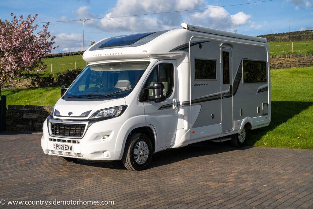 A white 2021 Auto-Sleepers Broadway EL motorhome is parked on a paved area with grassy surroundings and a blue sky with scattered clouds. The motorhome has tinted windows, an extended roof, and bears the number plate PO21 EKX. A tree in bloom and a stone wall are visible in the background.