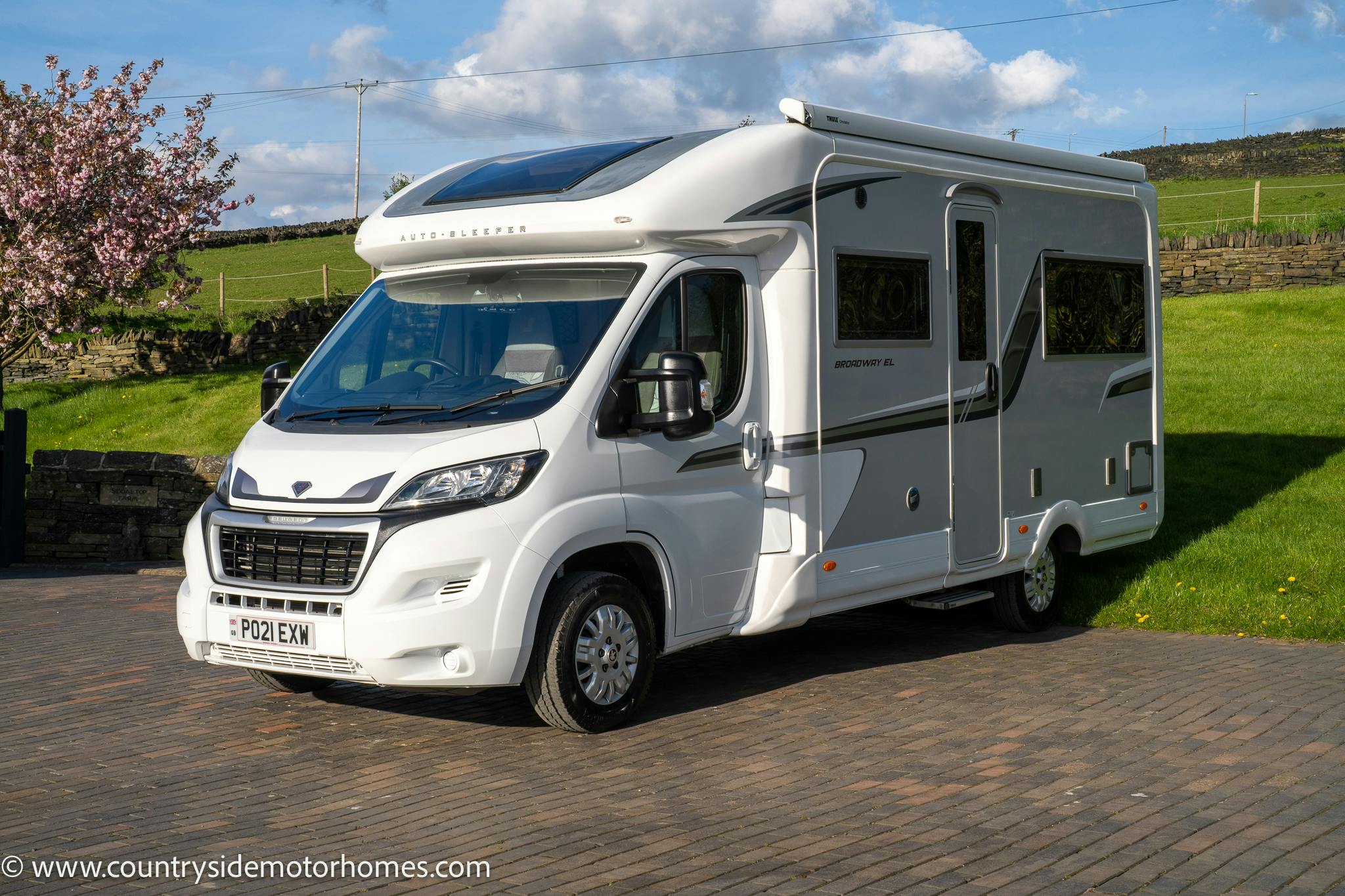 A modern white 2021 Auto-Sleepers Broadway EL motorhome with a UK license plate is parked on a paved area. The vehicle has large windows, a sleek design with grey accents, and solar panels on the roof. The background features a grassy area, blooming tree, stone wall, and blue sky with clouds.