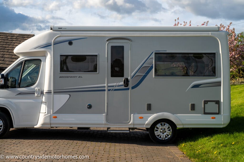 A 2021 Auto-Sleepers Broadway EL, with white bodywork and blue and grey accents, is parked on a brick driveway. The motorhome features a large side window, a door in the middle section, and smaller windows near the front and back. Green grass and trees are in the background.