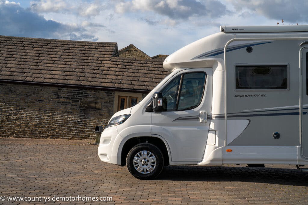 A white recreational vehicle, the 2021 Auto-Sleepers Broadway EL, is parked on a cobblestone driveway. There is a stone building with a dark shingled roof in the background. The RV proudly displays "Broadway EL" on its side. The sky is partly cloudy.