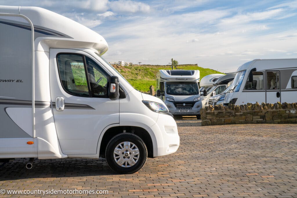 A paved lot with multiple parked motorhomes, including a prominent white 2021 Auto-Sleepers Broadway EL in the foreground. The background features other motorhomes, a stone wall, and a grassy hill under a partly cloudy sky. A website URL is seen at the bottom left.