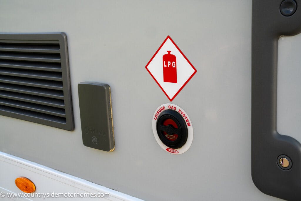 Close-up of a gray 2021 Auto-Sleepers Broadway EL exterior showing a ventilation grille, storage compartment, and a refill cap labeled for liquefied petroleum gas (LPG) with a red warning symbol above it. The website URL "www.countrysidemotorhomes.com" is visible at the bottom left.