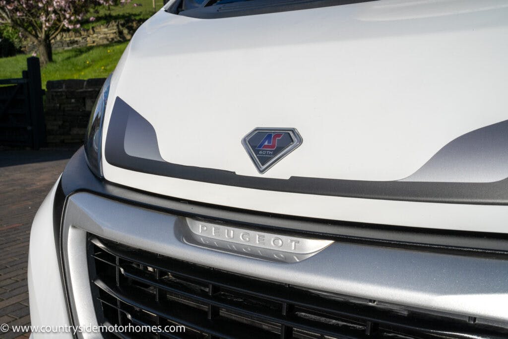 Close-up view of the front grille and emblem of a white Peugeot motorhome. The image shows a logo above the grille, featuring the text "Bolero" under the Peugeot brand name. This 2021 Auto-Sleepers Broadway EL model stands out with its sleek design against a blurred outdoor backdrop with greenery.