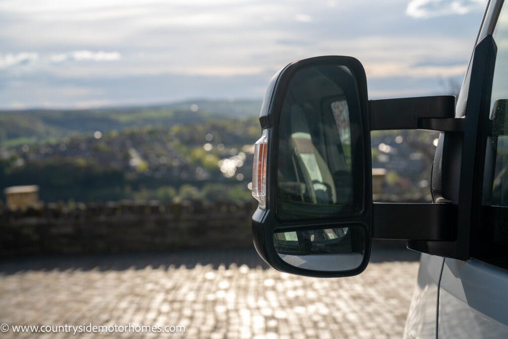 The image showcases a close-up of a 2021 Auto-Sleepers Broadway EL side mirror with a blurred background of hilly landscape and cloudy sky. The mirror reflects part of the vehicle and surrounding scenery, while the website URL "www.countrysidemotorhomes.com" is visible in the bottom left corner.