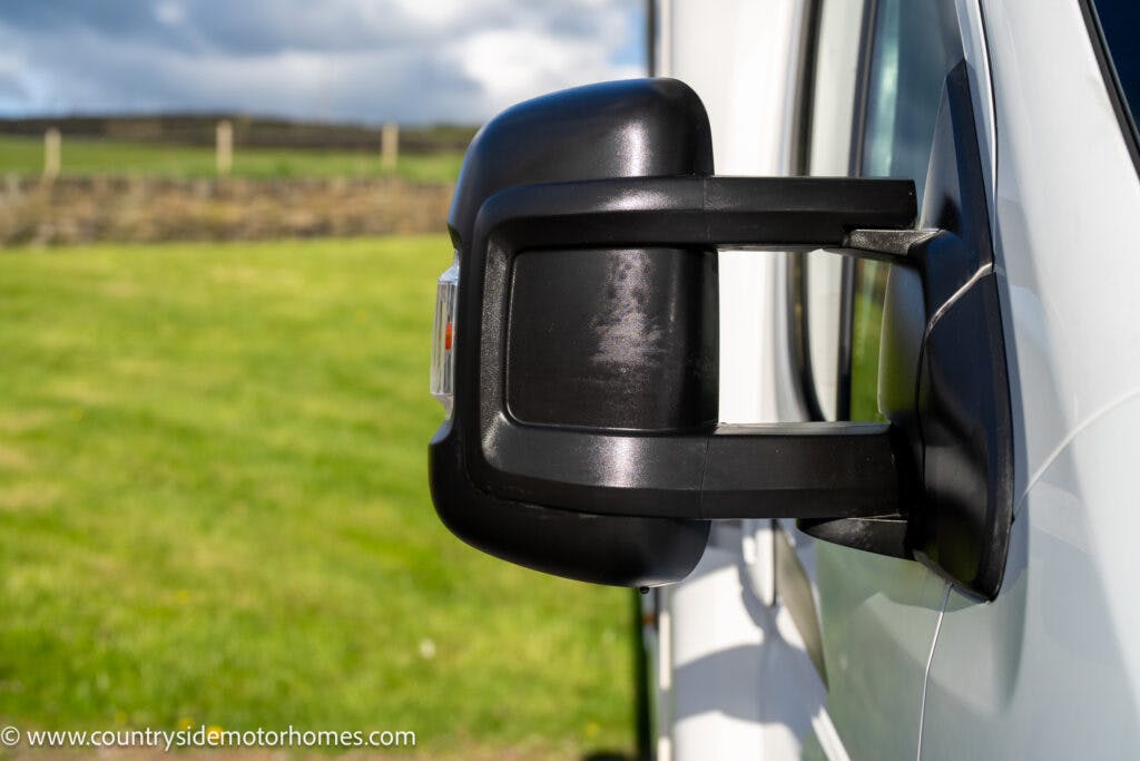 Close-up of a side mirror on a white 2021 Auto-Sleepers Broadway EL, with a grassy field and stone fence in the background. The sky is partly cloudy. The website "www.countrysidemotorhomes.com" is visible in the bottom left corner.