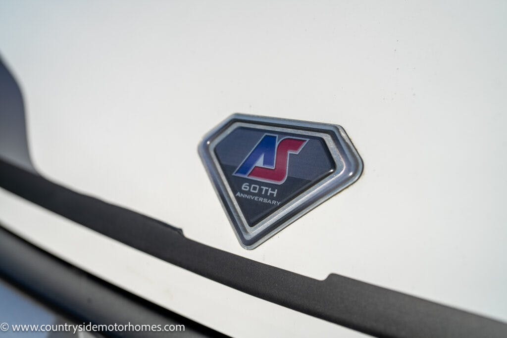 A close-up image of a vehicle’s emblem on a white surface. The emblem is diamond-shaped with "AS" in blue and red, and the text "60th Anniversary" below it. The website "www.countrysidemotorhomes.com" is visible in the lower left corner, showcasing the 2021 Auto-Sleepers Broadway EL.