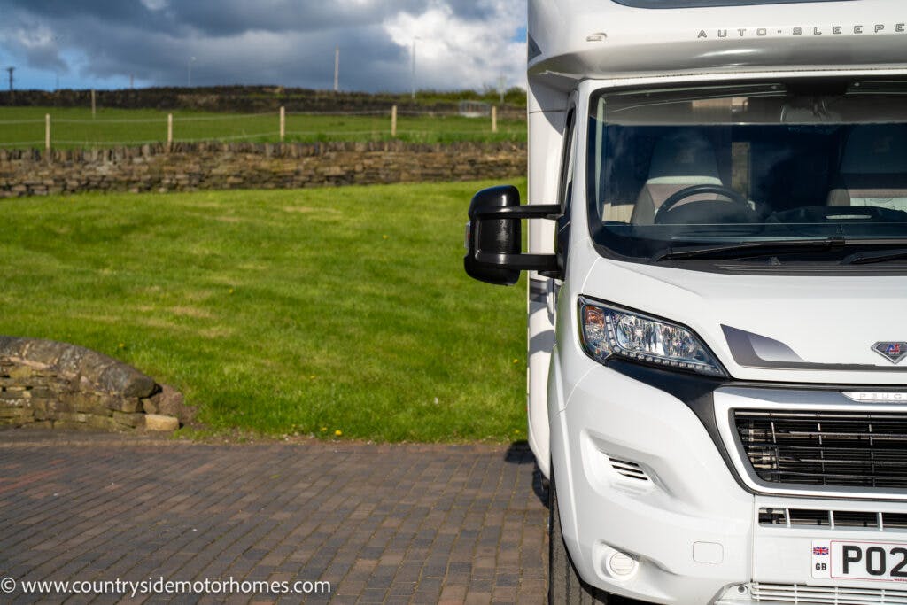 A white 2021 Auto-Sleepers Broadway EL camper van is parked on a paved area next to a grassy field with a stone wall in the background. The image has the website "www.countrysidemotorhomes.com" watermarked in the bottom left corner. The sky is partly cloudy.