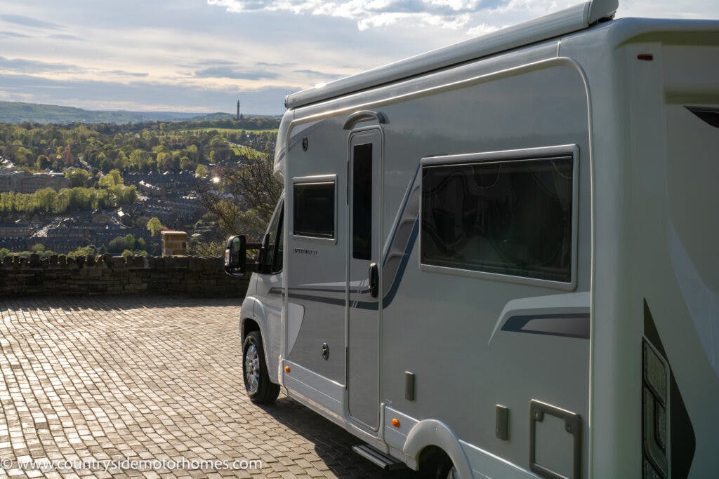 A 2021 Auto-Sleepers Broadway EL motorhome is parked on a paved area overlooking a scenic landscape. The background features rolling hills, a distant town, and a cloudy sky. The motorhome has a large side window and visible branding details.