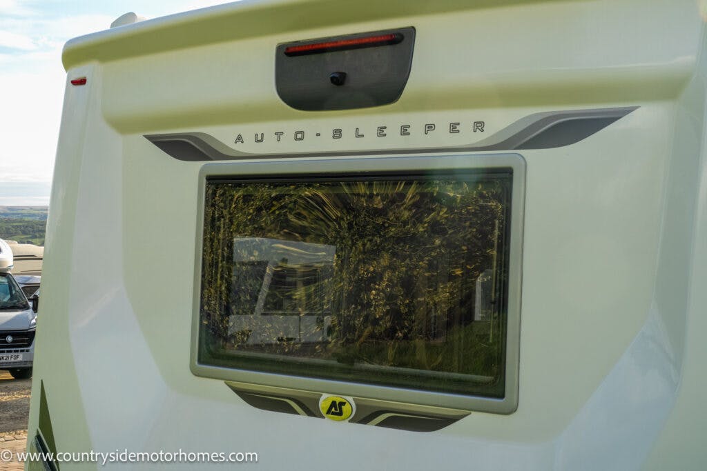 The image shows the rear view of a 2021 Auto-Sleepers Broadway EL motorhome. The back window is visible along with the motorhome's name displayed above it. The vehicle is cream-colored, and part of a website logo can be seen in the bottom left corner of the image.