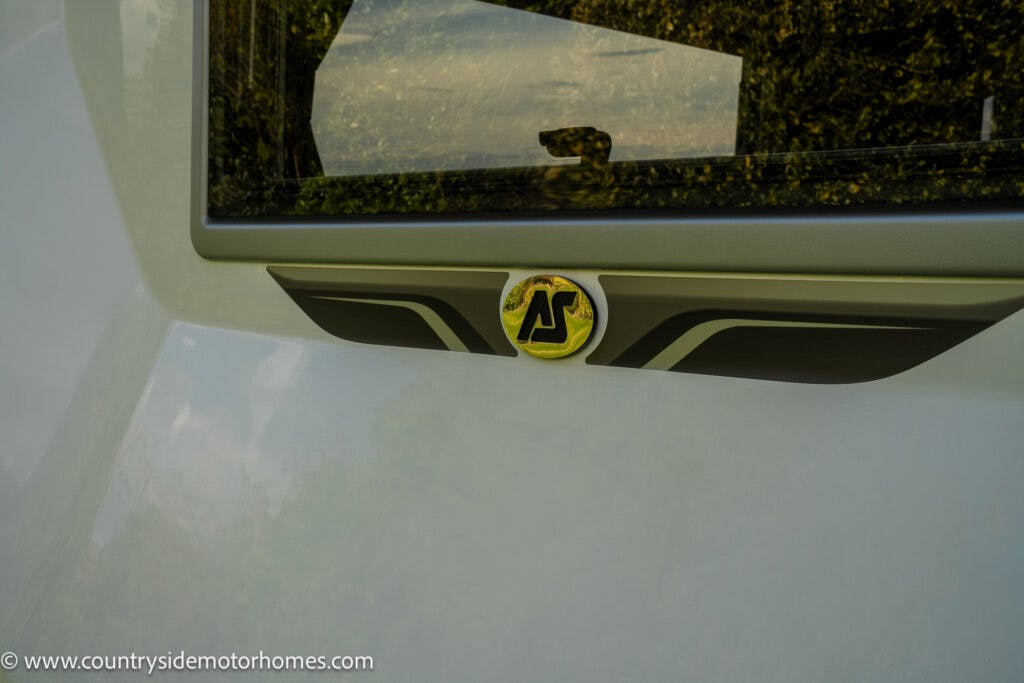 A close-up image of the rear window of a 2021 Auto-Sleepers Broadway EL. The lower portion of the window is framed by a decorative sticker with the initials "AS." The background shows some greenery reflected in the window. The website www.countrysidemotorhomes.com is visible in the corner.