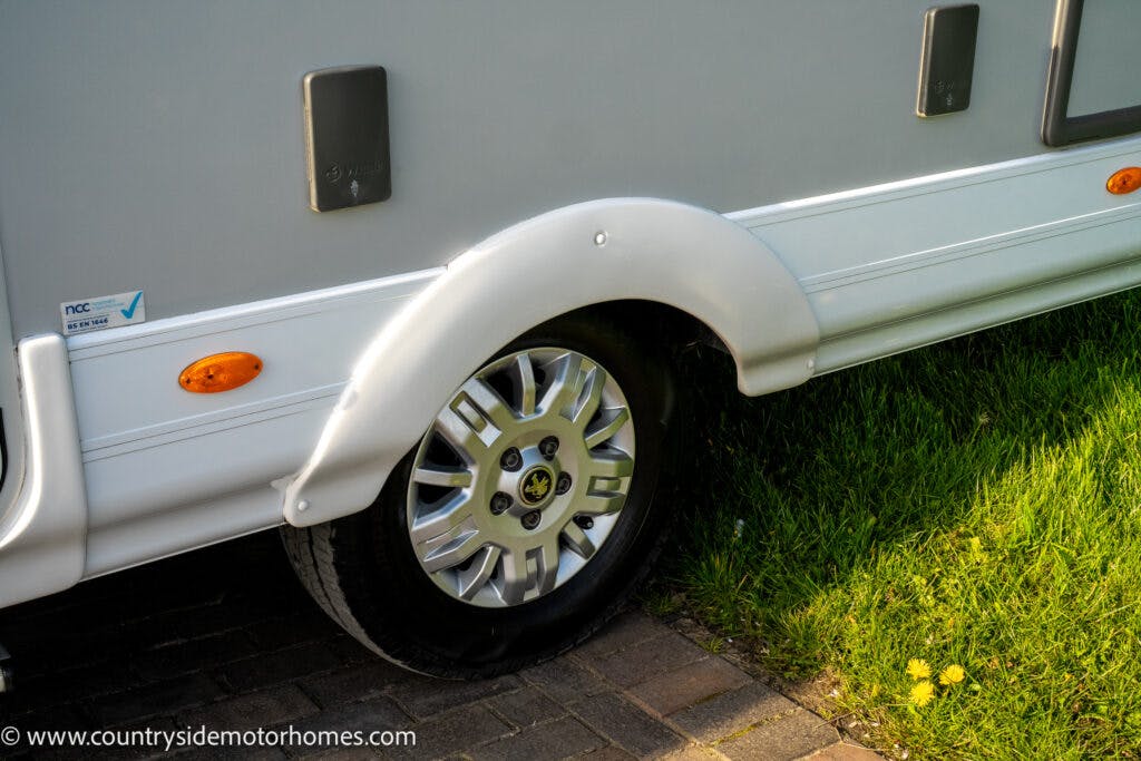 A close-up image of a 2021 Auto-Sleepers Broadway EL motorhome's wheel and side panel. The vehicle is parked on a combination of grass and a paved driveway. The web address www.countrysidemotorhomes.com is visible on the vehicle's exterior.