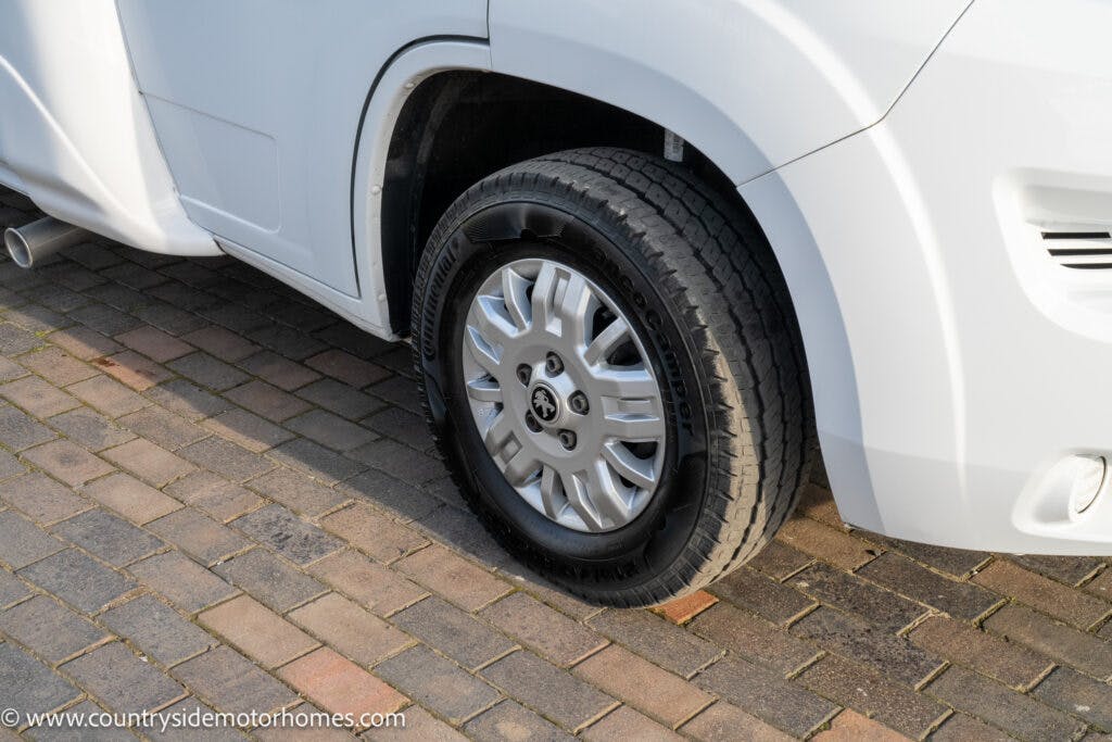 Close-up of the front tire and wheel of a white 2021 Auto-Sleepers Broadway EL parked on a brick driveway. The tire has a black sidewall, while the wheel is silver with a multi-spoke design. Part of the vehicle's bumper and side panel are visible.