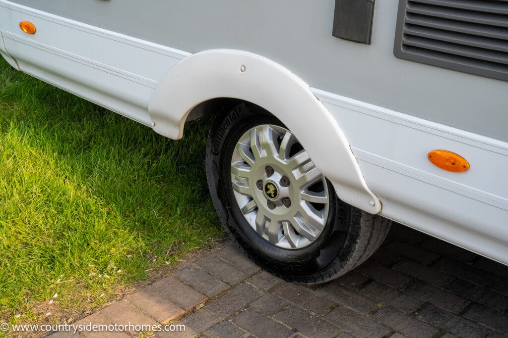 Close-up image of a 2021 Auto-Sleepers Broadway EL's rear wheel and tire, partially covered by a white fender. The RV is parked on a combination of grass and a brick driveway. The website URL "www.countrysidemotorhomes.com" is visible in the bottom left corner of the image.