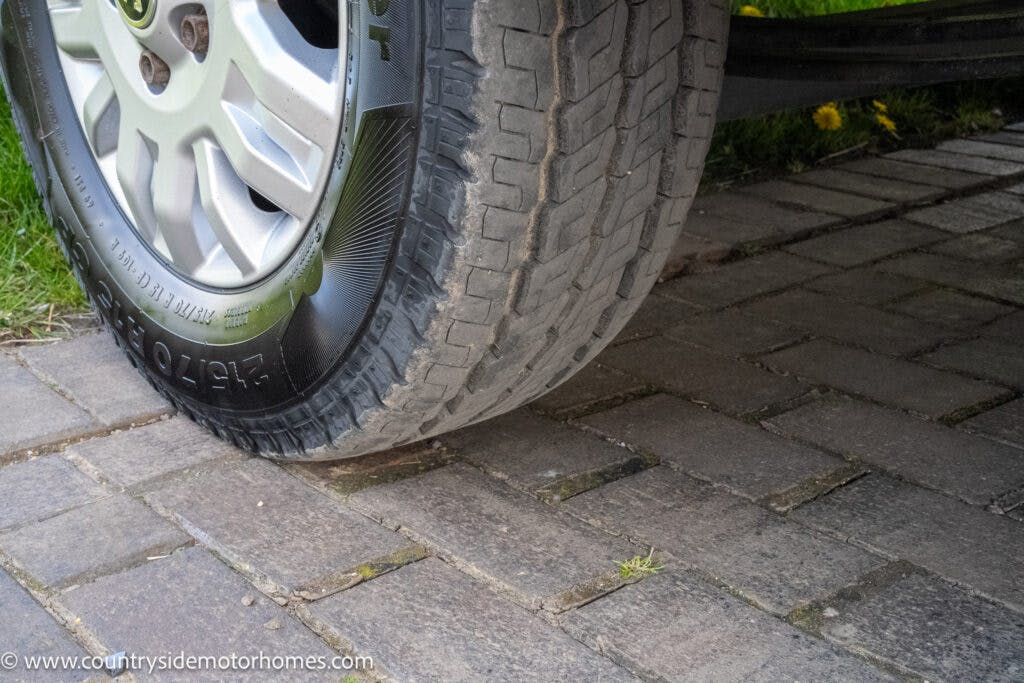 Close-up of a 2021 Auto-Sleepers Broadway EL's rear tire parked on a brick driveway. The tire appears to be in good condition with visible tread. The surrounding area includes some grass and a few dandelions. The image has a website watermark at the bottom left: www.countrysidemotorhomes.com.