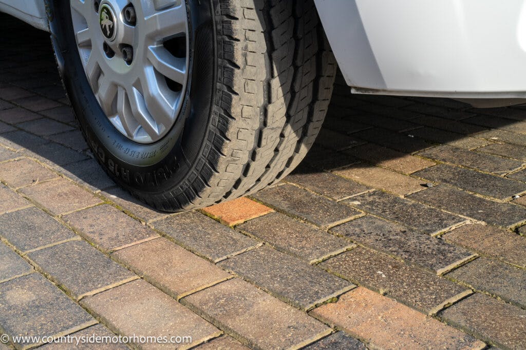 A close-up image shows the rear wheel of a 2021 Auto-Sleepers Broadway EL parked on a brick-paved surface. Sunlight casts shadows on part of the vehicle and the ground. The website www.countrysidemotorhomes.com is visible in the lower left corner.