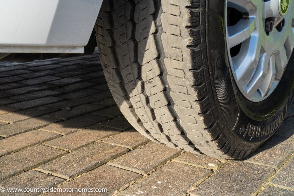 A close-up image of a 2021 Auto-Sleepers Broadway EL's front tire on a cobblestone driveway. The tire is partially worn, revealing the tread pattern. The rim is visible with five lug nuts. There is a website URL, www.countrysidemotorhomes.com, in the bottom left corner.