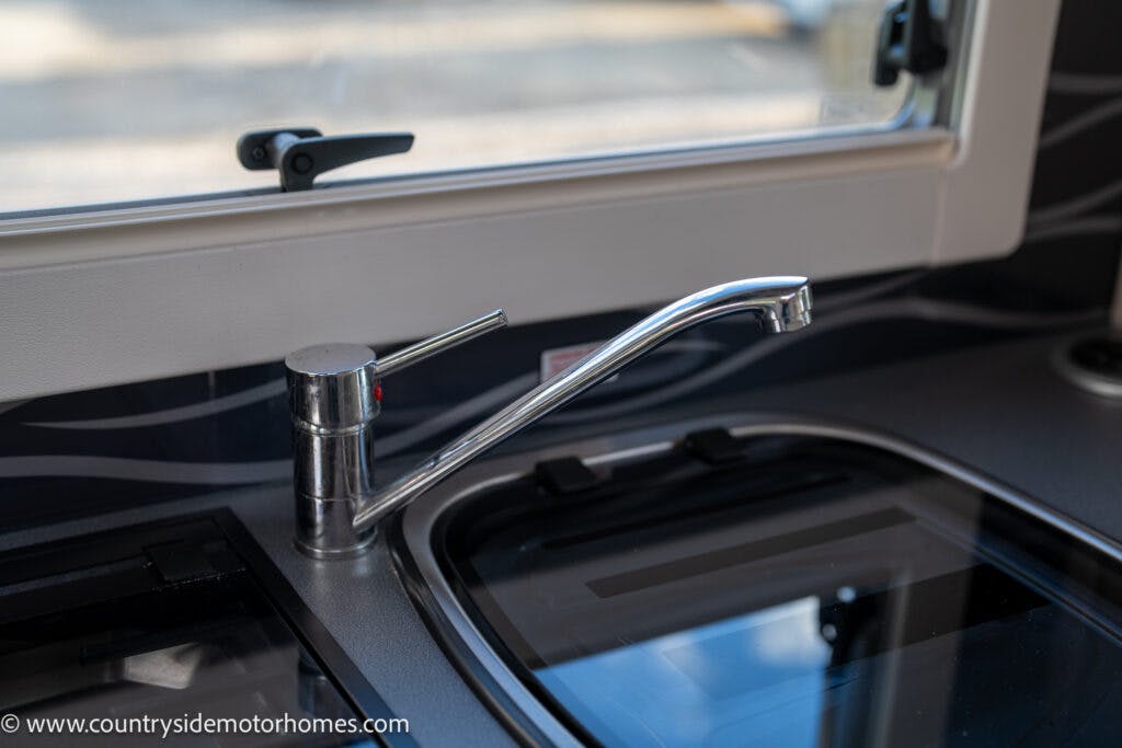 A close-up of a modern kitchen sink with a sleek, chrome faucet in the 2021 Auto-Sleepers Broadway EL. The sink is adjacent to a stovetop with a glass cover. The scene is well-lit, indicating a clean and well-maintained kitchen environment. A window above the sink allows natural light to enter.