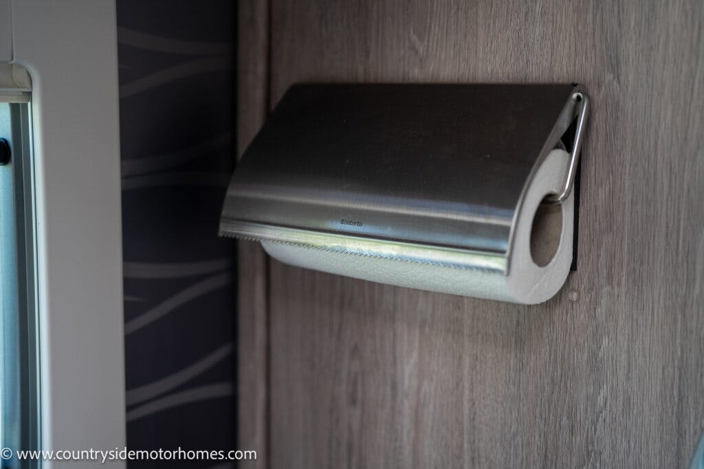 A metallic paper towel holder mounted on a wooden wall holds a roll of paper towels. Its sleek and modern design complements the high-end interiors of the 2021 Auto-Sleepers Broadway EL perfectly.