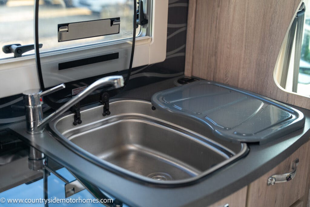 A compact kitchen sink setup inside a 2021 Auto-Sleepers Broadway EL motor home. The sink features a single faucet, a stainless steel basin, and a fitted lid cover. Surrounding the sink are countertop surfaces and a window above the sink. A website URL is visible in the bottom-left corner.