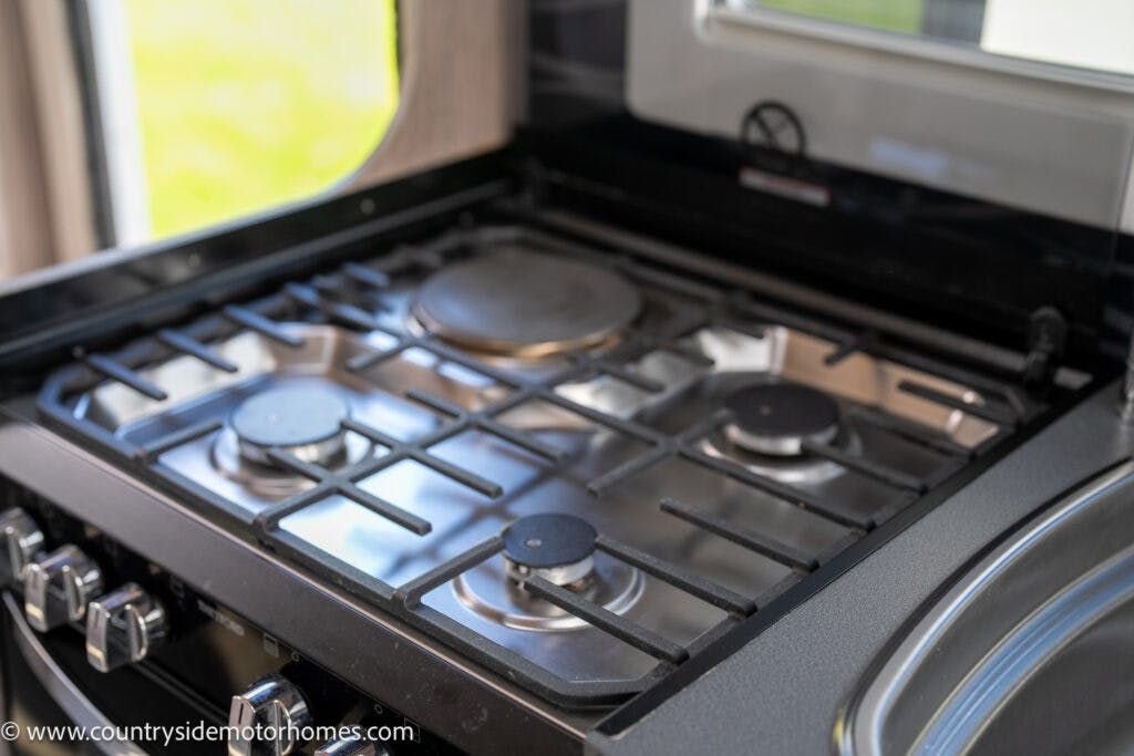 A sleek 2021 Auto-Sleepers Broadway EL motorhome kitchen features a stainless steel four-burner gas stove with black grates, complemented by side control knobs and positioned next to a metal sink. A window is partially visible in the background.
