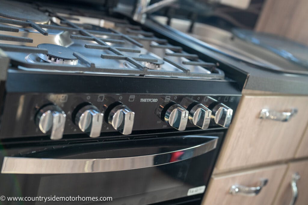 Close-up of a modern kitchen stove with five control knobs and a digital display in the 2021 Auto-Sleepers Broadway EL. The stove boasts a stainless steel gas cooktop and an oven below, with a portion of the sink and wooden cabinetry visible in the background.