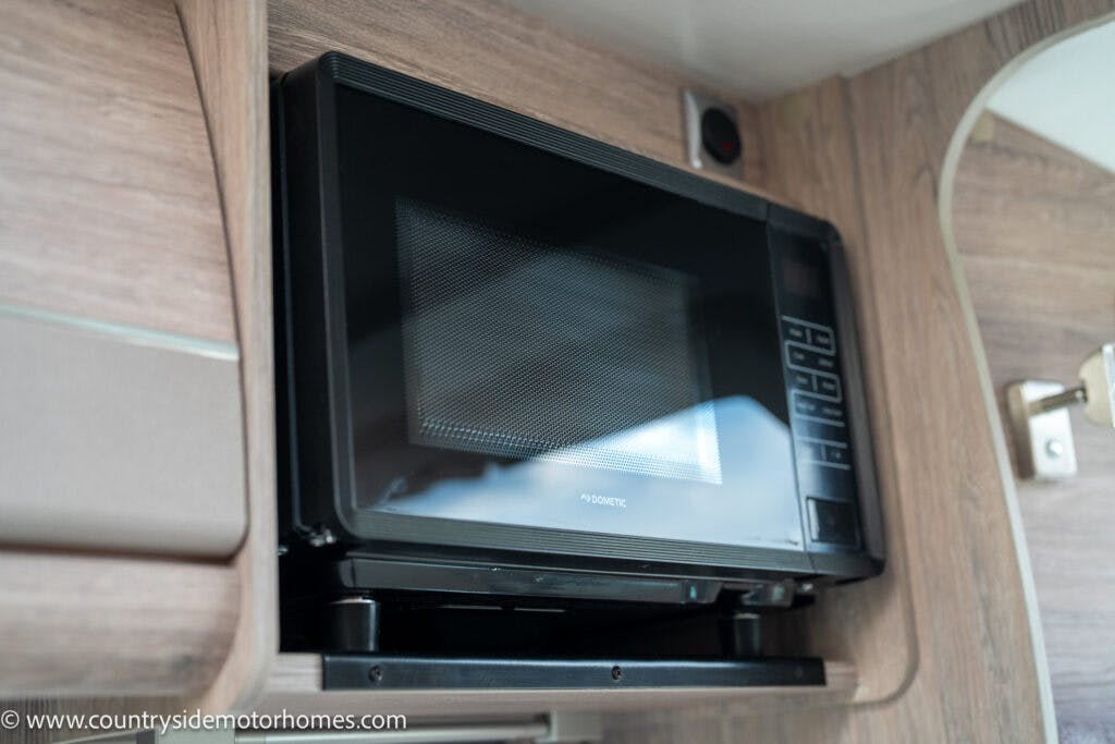 A black microwave is installed in a wooden cabinet inside the 2021 Auto-Sleepers Broadway EL. The microwave has a digital keypad and display on its right side. The light wood grain of the cabinet complements the motorhome's interior, and the door of the microwave is closed.