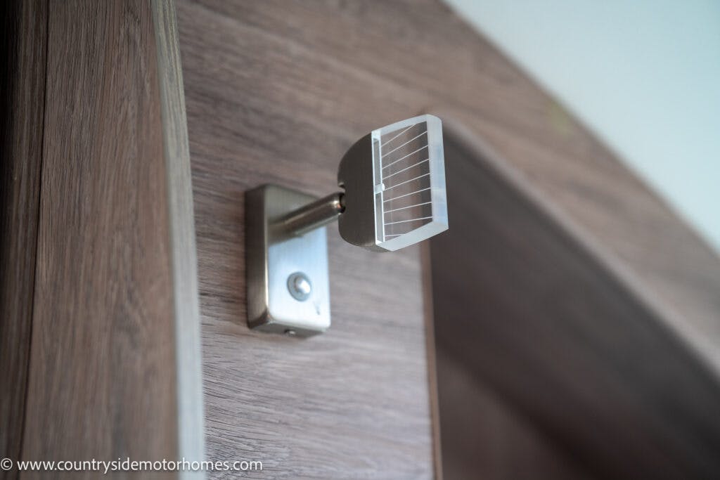 A close-up of a wall-mounted modern light fixture with a chrome base and a clear geometric lampshade, positioned against a wood-paneled wall in the 2021 Auto-Sleepers Broadway EL. The fixture is turned off. The website "www.countrysidemotorhomes.com" is visible in the bottom left corner.