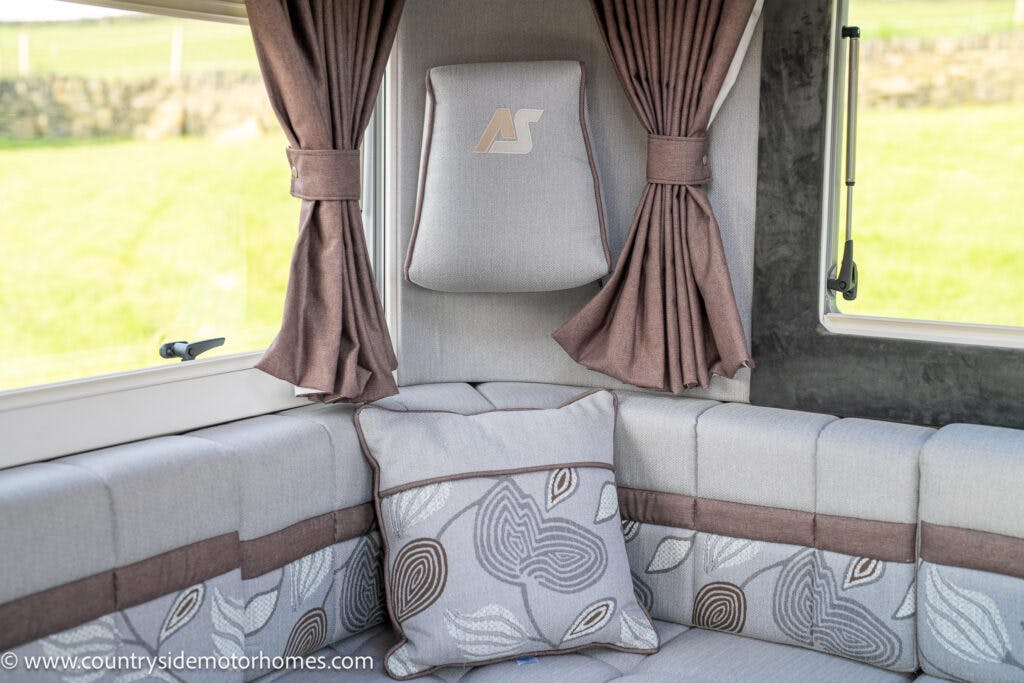 Interior of a 2021 Auto-Sleepers Broadway EL motorhome with a gray and brown color scheme. The image shows cushioned seating with patterned pillows, a headrest featuring an "AS" logo, and brown curtains tied back. A window offers a view of the green landscape outside.