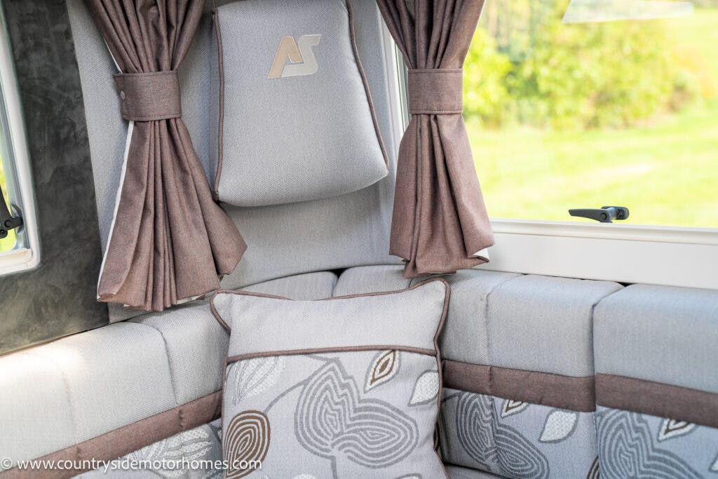 The image shows the corner of a 2021 Auto-Sleepers Broadway EL motorhome seating area with grey upholstered cushions and a matching pillow featuring a leaf pattern. Brown curtains are drawn to the sides of a window that provides a view of green outdoors. A logo is visible on the headrest.