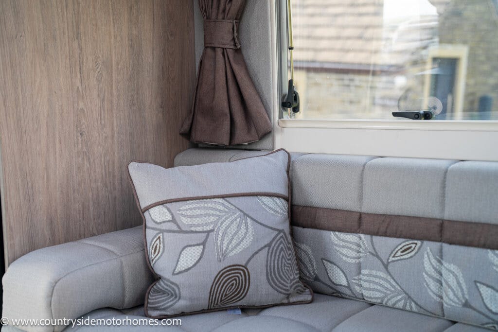A close-up of the 2021 Auto-Sleepers Broadway EL interior shows a gray upholstered couch with a matching pillow featuring a leaf pattern. Behind the couch is a window with a drawn brown curtain. The website www.countrysidemotorhomes.com is visible in the bottom left corner.