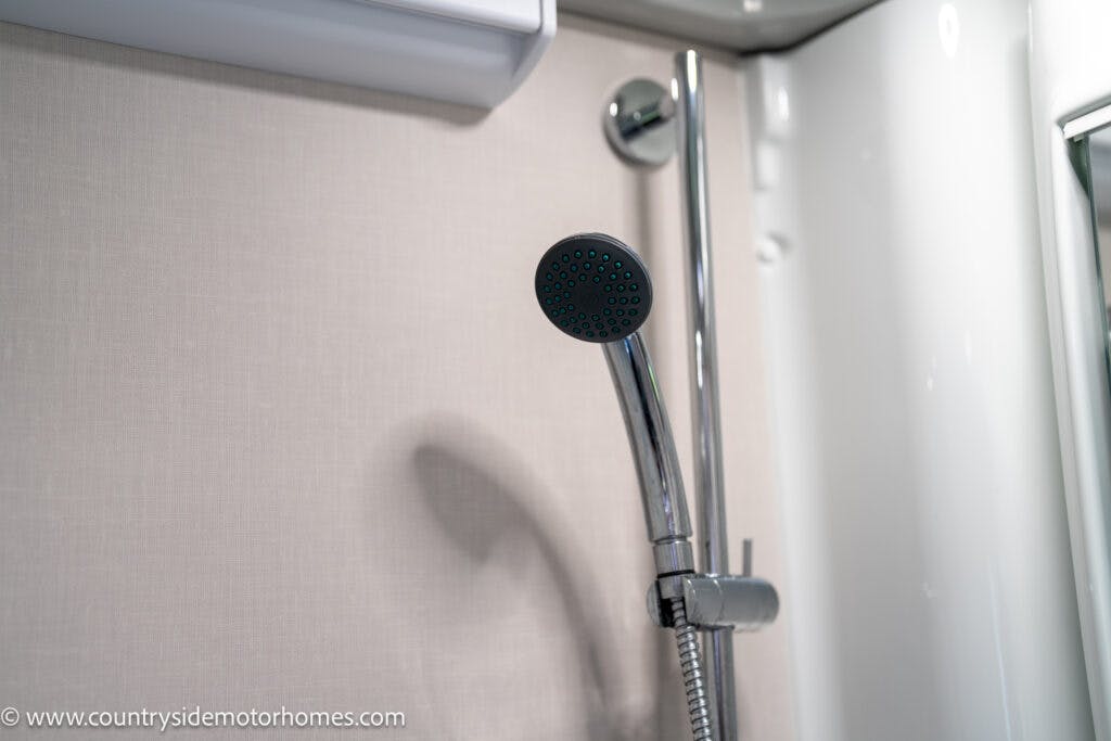 A close-up view of a modern shower head attached to a wall in the bathroom of a 2021 Auto-Sleepers Broadway EL. The metallic shower head with a flexible hose is mounted near a chrome handle, set against a light-colored wall background.