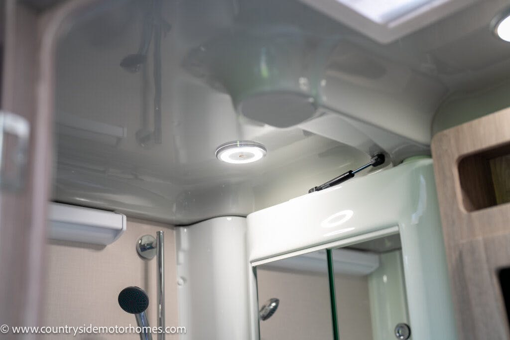 Interior view of the 2021 Auto-Sleepers Broadway EL motorhome bathroom highlighting a shiny surface ceiling with embedded LED lights, a wall-mounted showerhead, a bathroom mirror, and a window with a vent. The reflection in the mirror shows additional bathroom details.