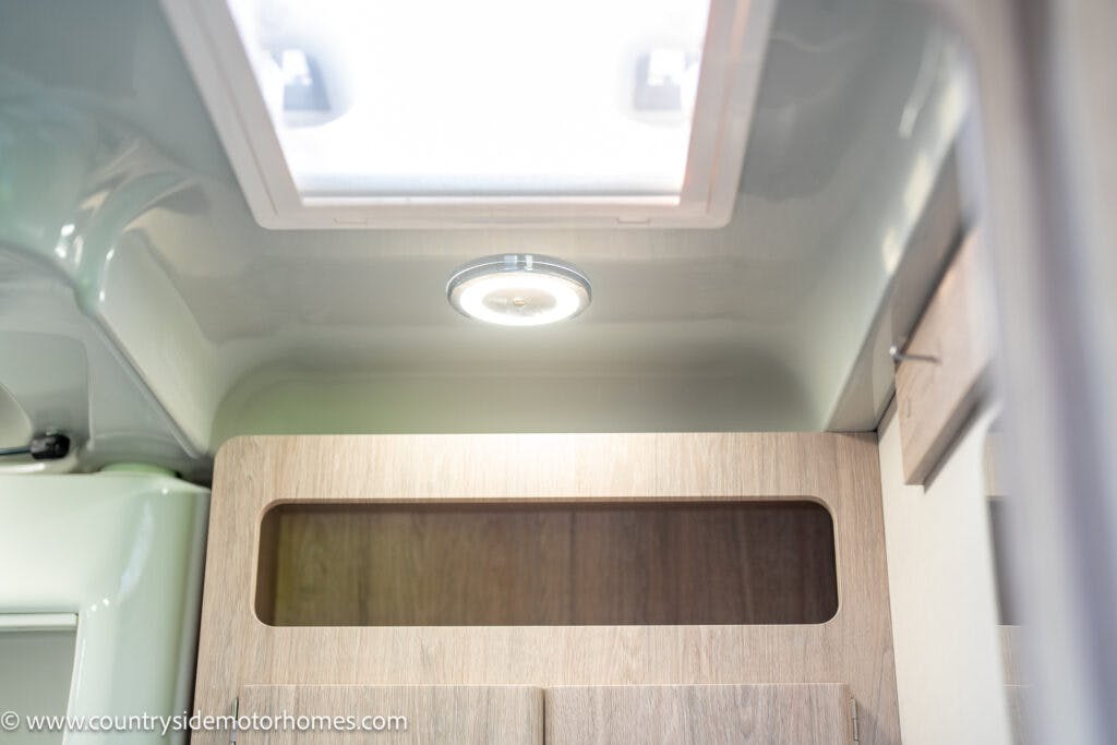 The image showcases the interior roof of a 2021 Auto-Sleepers Broadway EL motorhome, highlighting a ceiling light and a skylight. Below them, there is wooden cabinetry with an open compartment. The photo is taken from a low angle, looking upward.