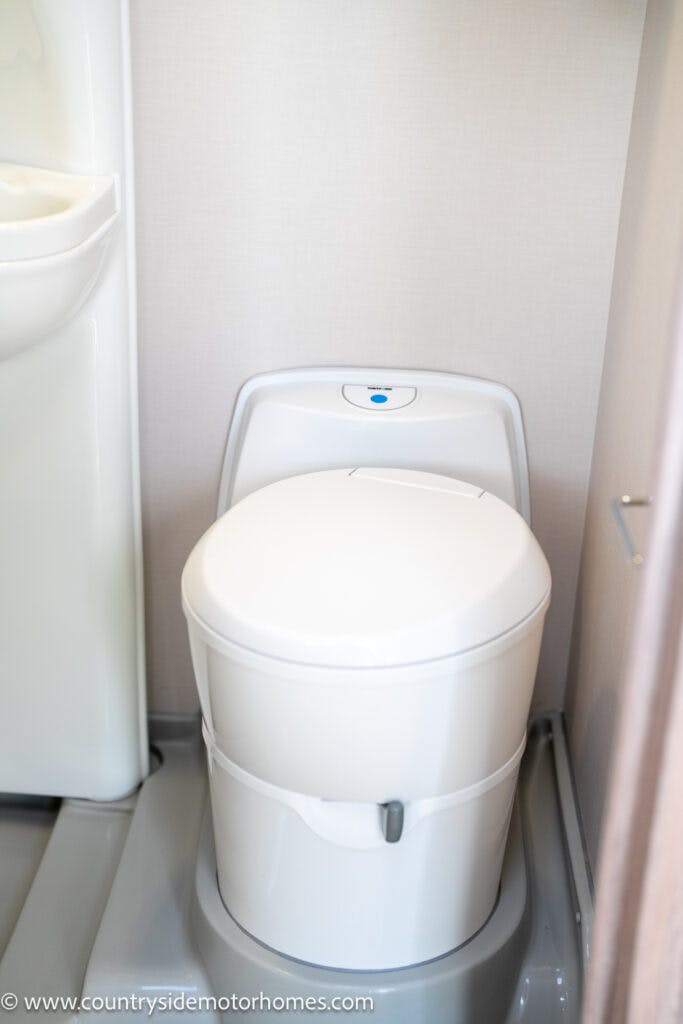 A white compact portable toilet, similar to those found in the 2021 Auto-Sleepers Broadway EL, is positioned in a small corner with a minimalistic design and a small control button on top. A part of a white sink is visible to the left side of the image. The surroundings are clean and simple.