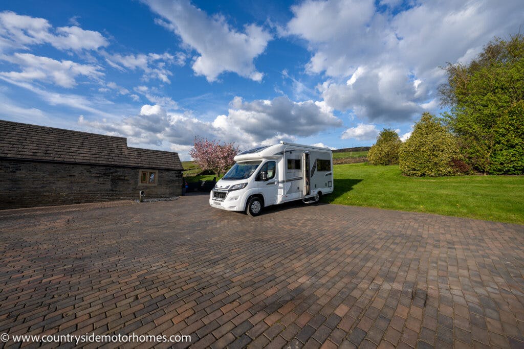 A white 2021 Auto-Sleepers Broadway EL motorhome is parked on a large, paved driveway surrounded by green lawn, trees, and buildings. A cloud-dotted blue sky is visible above. The URL "www.countrysidemotorhomes.com" can be seen on the bottom left corner of the image.