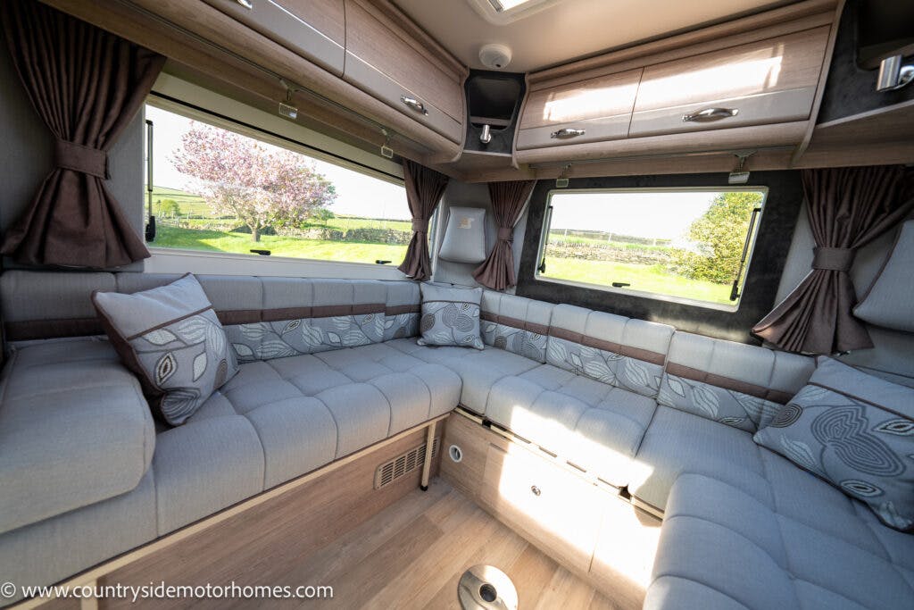The image shows the interior of a 2021 Auto-Sleepers Broadway EL motorhome with a spacious seating area. The seating is upholstered in light gray fabric with patterned pillows. There are windows with brown curtains and wooden cabinets above the seats. Sunlight illuminates the space.