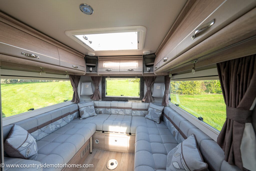 The image shows the interior of a 2021 Auto-Sleepers Broadway EL motorhome with an ample seating area upholstered in light grey fabric. The space includes overhead storage cabinets, large windows with curtains, and a central skylight. The floor appears to be made of light wood.