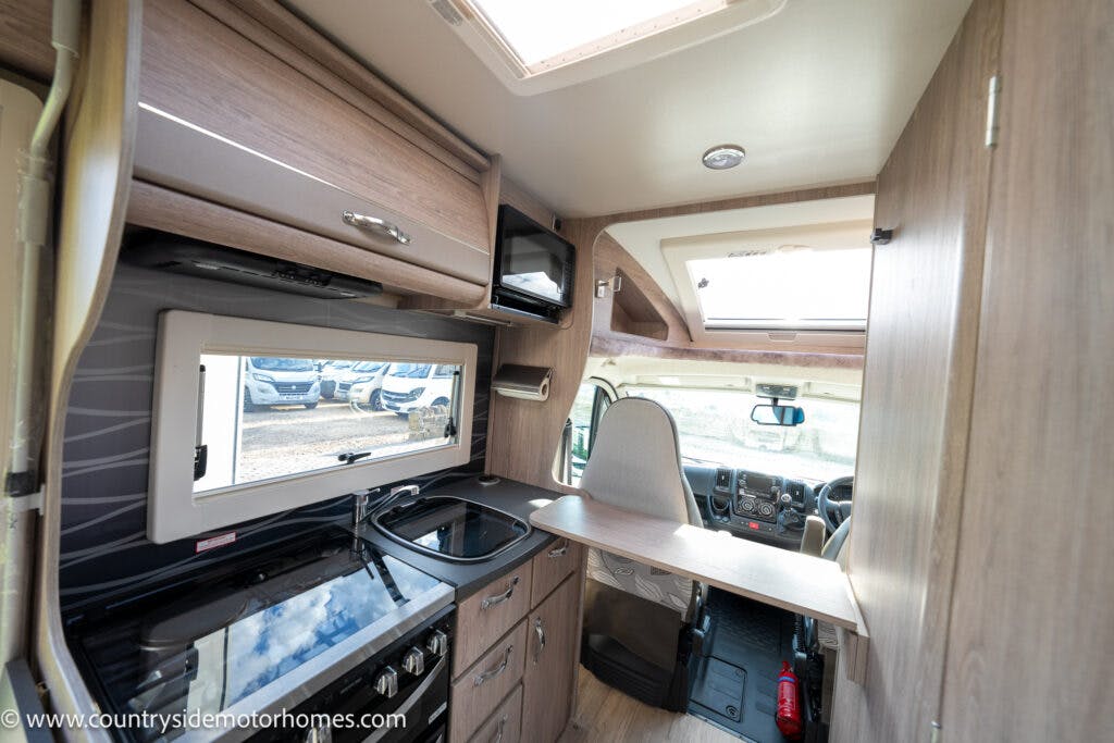 Interior view of a 2021 Auto-Sleepers Broadway EL motorhome showing a compact kitchen area with a sink, stove, and microwave. Overhead cabinets provide storage. The view extends to the driver's cockpit area, which features a steering wheel and dashboard.