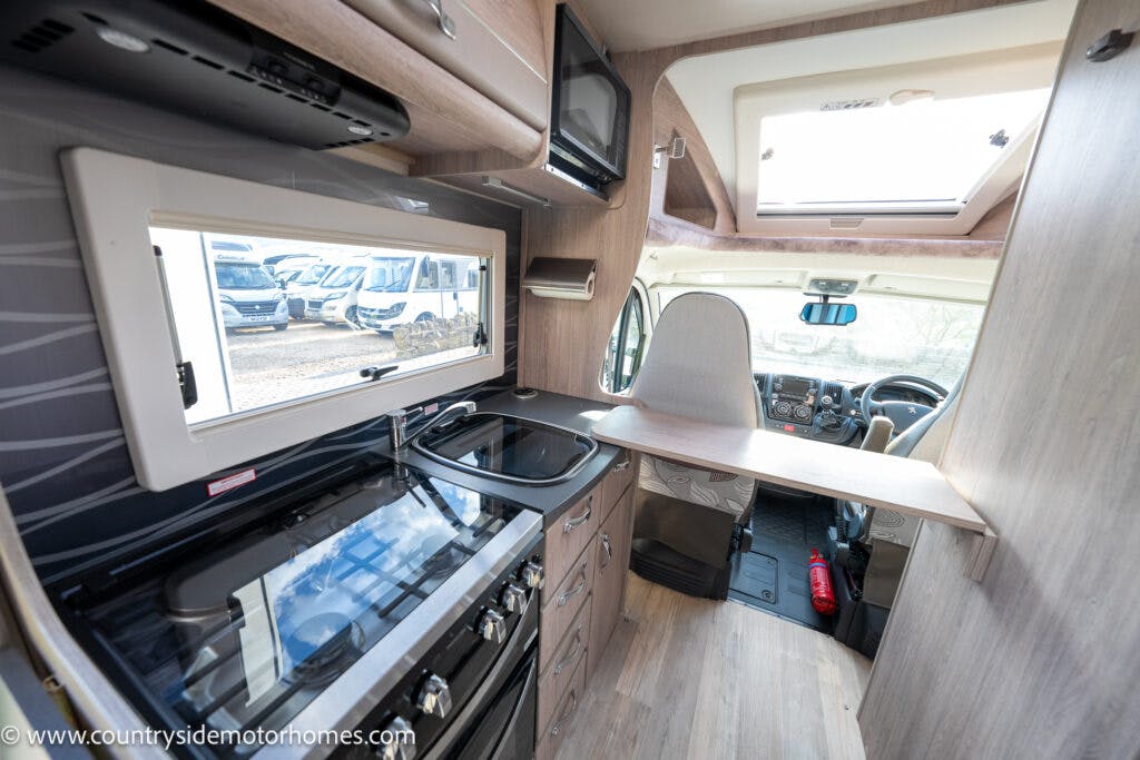 Interior of a 2021 Auto-Sleepers Broadway EL motorhome featuring a small kitchen area with a stove, sink, and countertop. A driver's seat is visible in the background. There's a rooftop window providing natural light, and several motorhomes are seen through the side window.