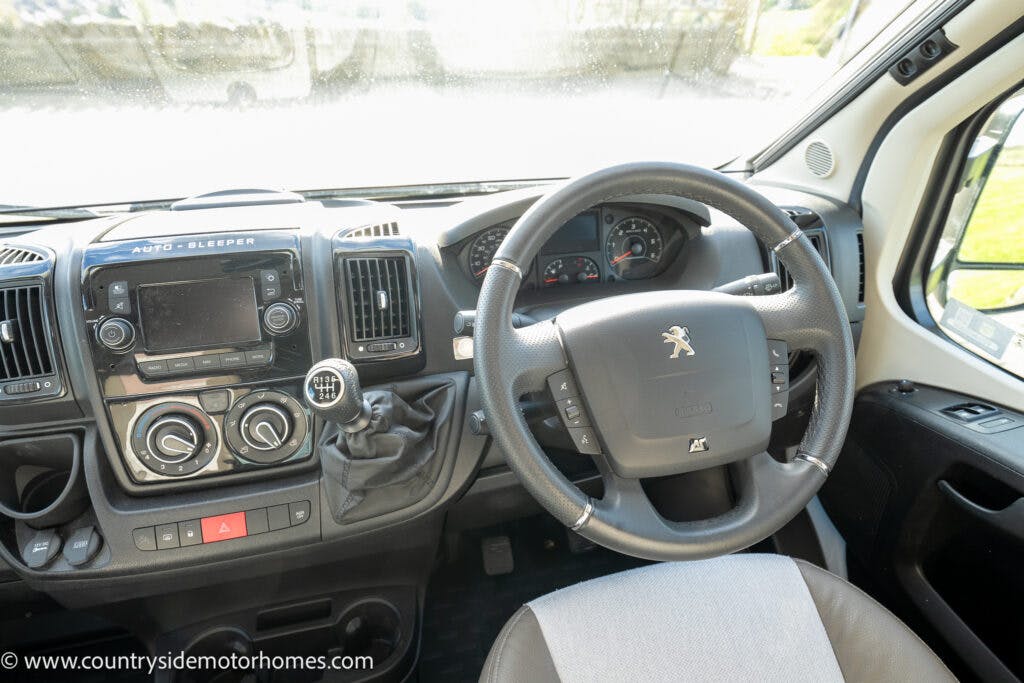 Interior view of a vehicle dashboard featuring a steering wheel with the Peugeot logo, control buttons, an infotainment screen, air vents, climate control knobs, and other standard components. The website www.countrysidemotorhomes.com is visible in the image of this 2021 Auto-Sleepers Broadway EL.