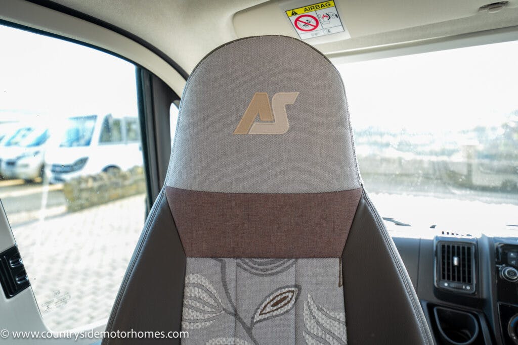 Close-up view of a car seat with "AS" embroidered on the headrest in a 2021 Auto-Sleepers Broadway EL. The upholstery is a mix of light gray and dark brown fabric with a leaf pattern on the bottom section. The dashboard and steering wheel are partially visible in the background.