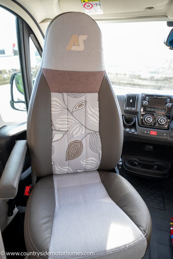 The image shows a driver's seat in a 2021 Auto-Sleepers Broadway EL motorhome. The seat is a combination of gray leather and patterned fabric with a mix of leaf designs. The dashboard and steering wheel are visible in the background, and a logo with the initials "AS" is displayed on the headrest.