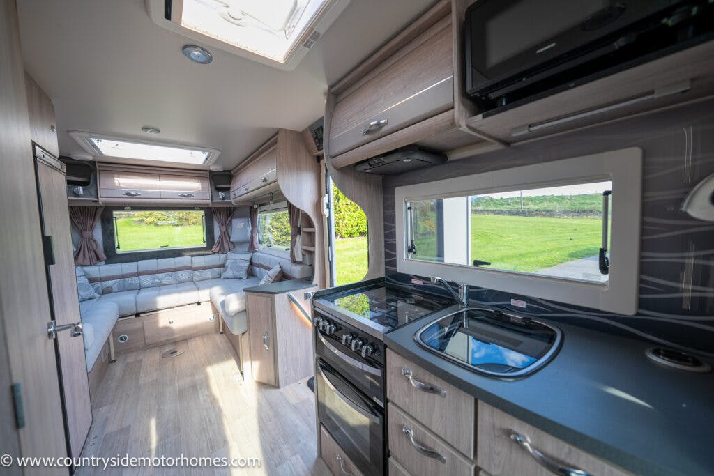 The image shows the interior of a 2021 Auto-Sleepers Broadway EL motorhome with wooden cabinets, a black stove with oven, black microwave, stainless steel sink, and a seating area with windows in the background. A green lawn can be seen through the motorhome's windows.