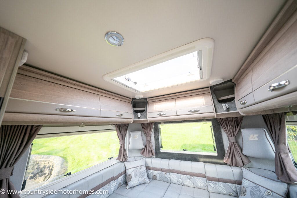 Interior of a 2021 Auto-Sleepers Broadway EL motorhome featuring a seating area with cushioned benches, overhead cabinets, brown curtains, and a skylight. The space is well-lit with natural light from the windows and skylight. "www.countrysidemotorhomes.com" is visible in the bottom left corner.