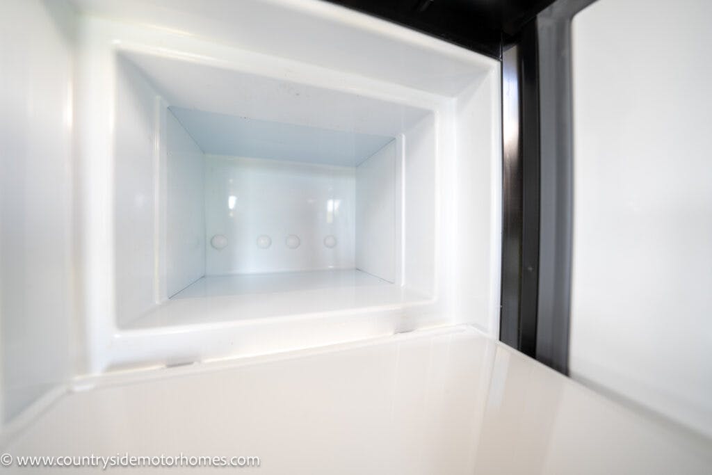 Close-up image of an empty white interior of a freezer compartment, surrounded by white plastic walls and a black door frame. The inside appears clean and well-lit, with the structure of the freezer clearly visible. Perfect for 2021 Auto-Sleepers Broadway EL adventures. The website URL is displayed in the bottom-left corner.